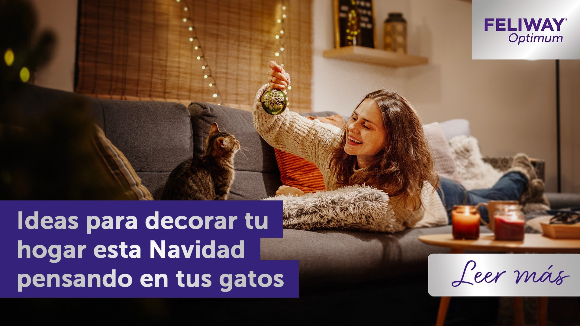 Mujer sonriente mostrando un adorno navideño brillante a un gato atigrado.
